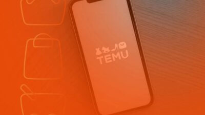 Smartphone sobre uma superfície de madeira exibindo o logotipo do aplicativo de compras Temu na tela, sob um filtro alaranjado.